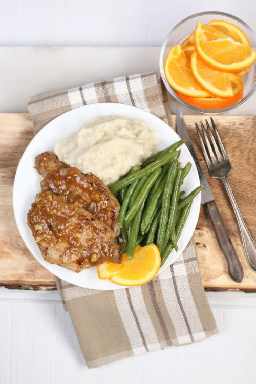 orange glazed pork chops on white plate with fresh green beans and mashed potatoes, orange slices in clear bowl to right