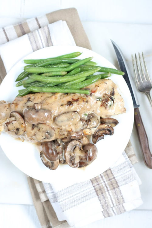 stuffed chicken breast with mushroom cream sauce on white plate with fresh green beans, fork and knife to right