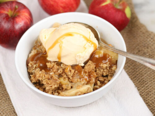 apple crisp in white bowl, topped with vanilla ice cream and drizzled with caramel sauce