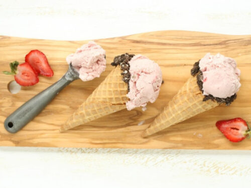ice cream cones with strawberry ice cream on wooden cutting board, vintage metal ice cream scoop to left