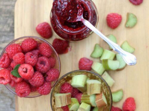 raspberry rhubarb jam in glass jar, spoon on top, on wooden cutting board, loose raspberries and rhubarb pieces