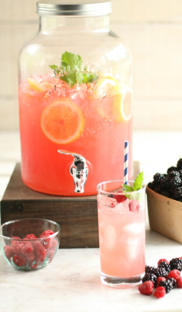 glass drink dispenser filled with lemonade, fresh lemon slices, fresh mint. Clear cylinder glass of lemonade in front with fresh berries, blue and white striped straw