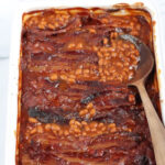baked beans topped with crispy bacon in white rectangle baking dish, wooden spoon in right