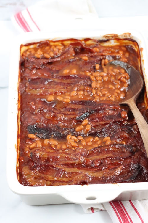 white rectangle baking dish with baked beans topped with crispy bacon, wooden spoon in right side of dish