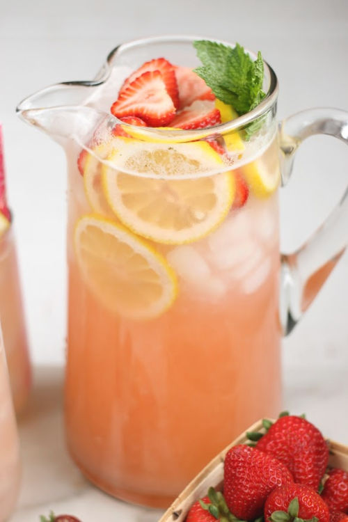 Clear glass pouring pitcher with strawberry lemonade, lemon slices, strawberry slices, and a sprig of fresh mint.