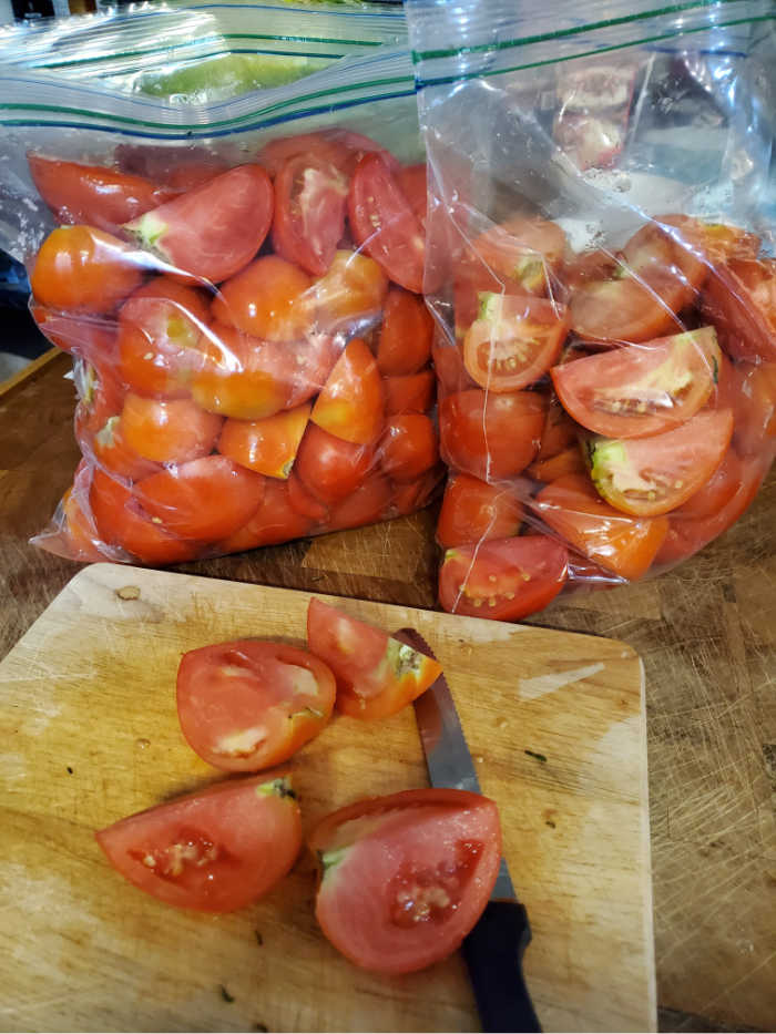 slicing fresh tomatoes on butcher block. Tomato quarters in two large zip-style freezer bags behind cutting board.