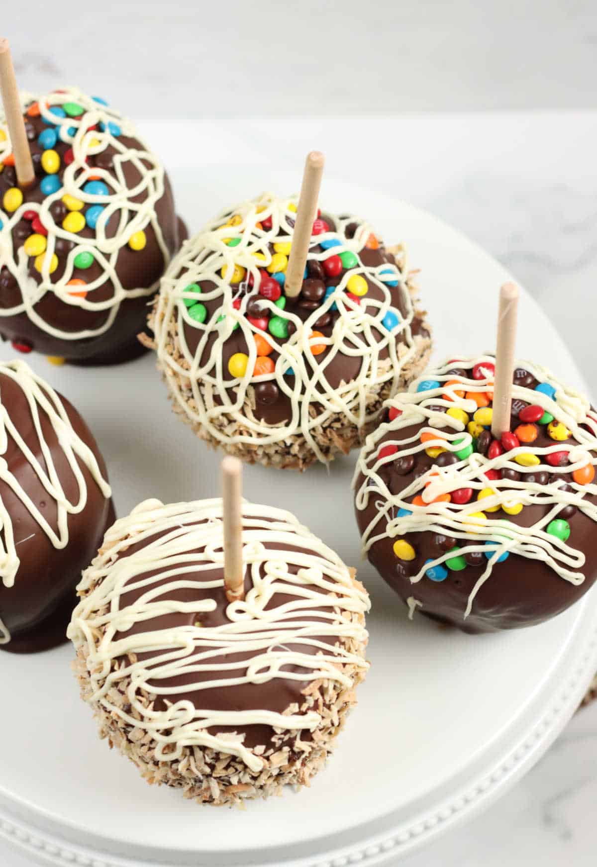 Caramel and chocolate dipped apples with toppings on white footed cake dish.