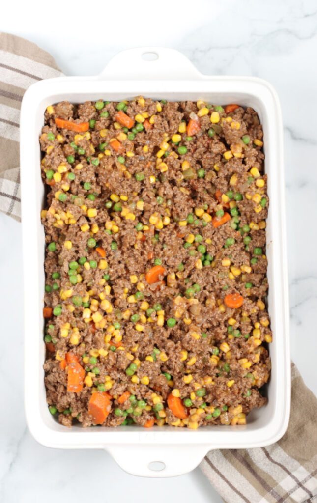 cooked ground beef, carrots, peas, corn, and onions in a white ceramic rectangle casserole dish, sitting on taupe plaid kitchen cloth