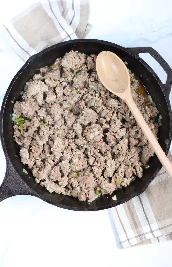 large cast iron skillet with browned meat, onions, and green peppers. Large spoon in pan.