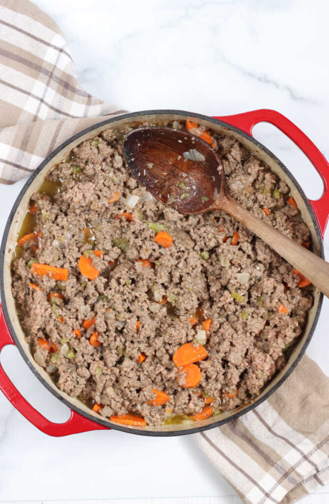 red enamelware pot with browned beef, carrots, onions, and celery pieces. Wooden spoon in the right of the pot.