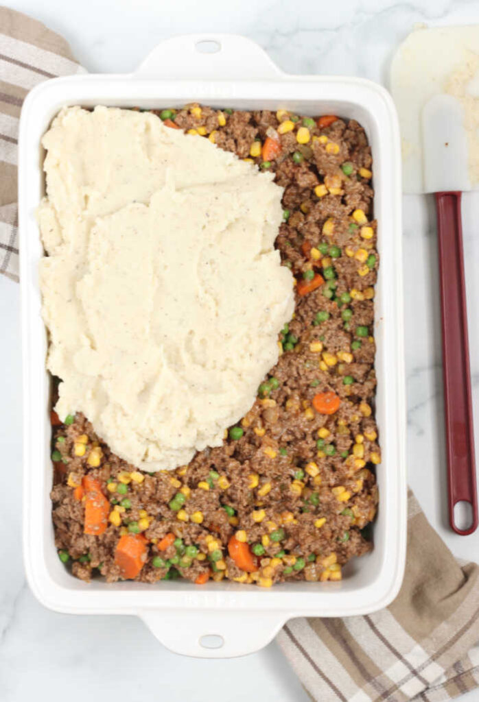 white ceramic rectangle shaped baking dish with shepherds pie, spreading the top layer of mashed potatoes