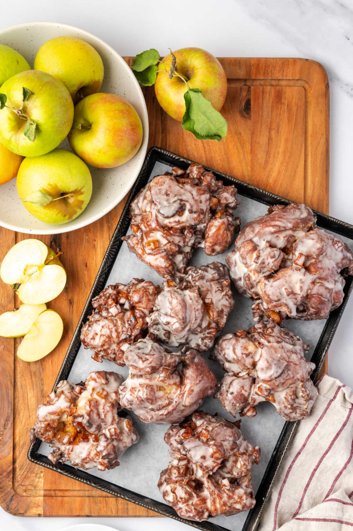 Apple fritters drying on sheet pan, apples in bowl on wood cutting board.
