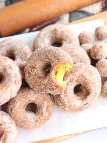 pumpkin donuts stacked on each other on wooden cutting board lined with white parchment paper, bite out of one donut