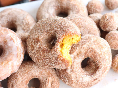 pumpkin donuts stacked on each other on wooden cutting board lined with white parchment paper, bite out of one donut