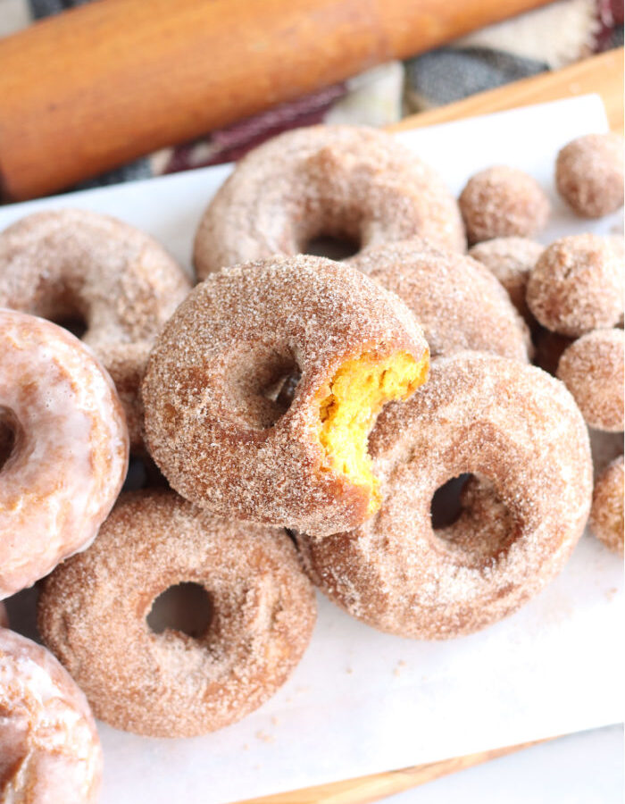 pumpkin donuts stacked on each other on wooden cutting board lined with white parchment paper, bite out of one donut