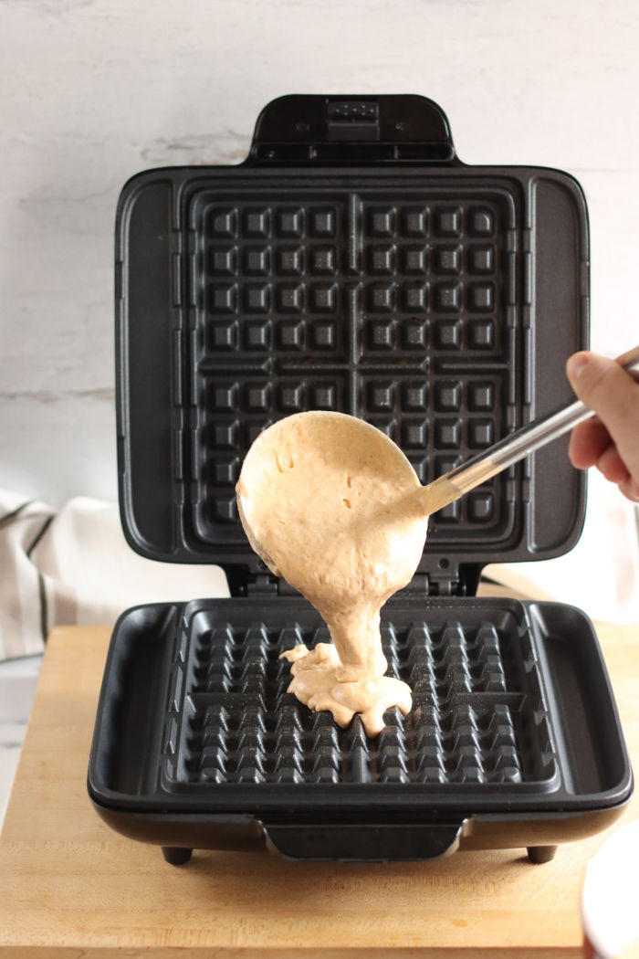Hand pouring pumpkin waffle batter with a metal scoop into a square waffle maker on a wooden cutting board 