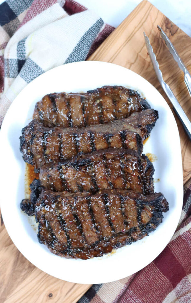 Four marinated steaks with grill marks on a oval white serving dish, metal tongs to the right