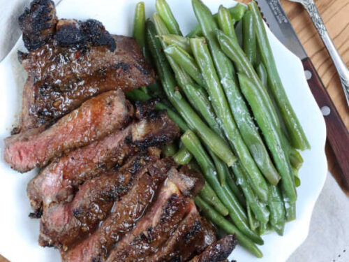 grilled steak sliced on small white plate, fresh green beans to the side of steak. Steak knife and fork next to plate on wooden cutting board