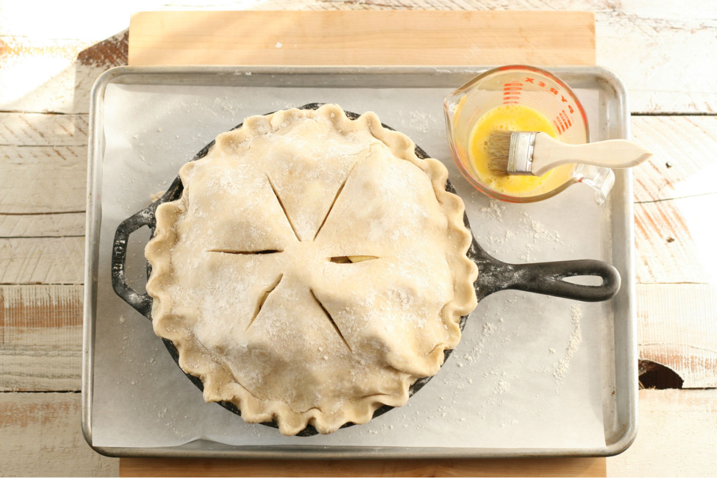 Apple pie in cast iron skillet on parchment lined half sheet pan, egg wash in glass measuring cup.