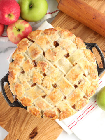Apple pie with lattice crust in cast iron pie plate, sitting on wooden cutting board.