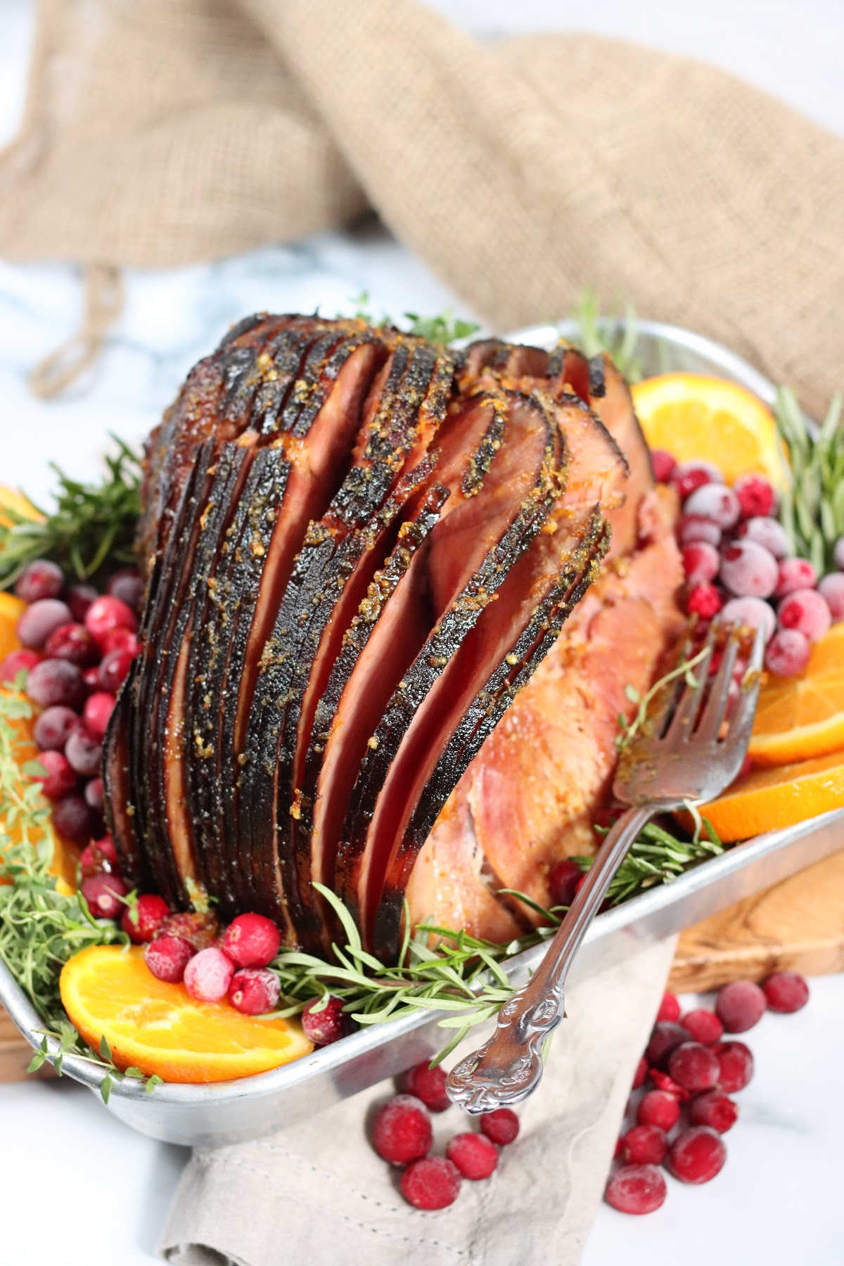 Spiral cut ham in metal baking pan, orange slices, fresh cranberries, fresh thyme and rosemary.