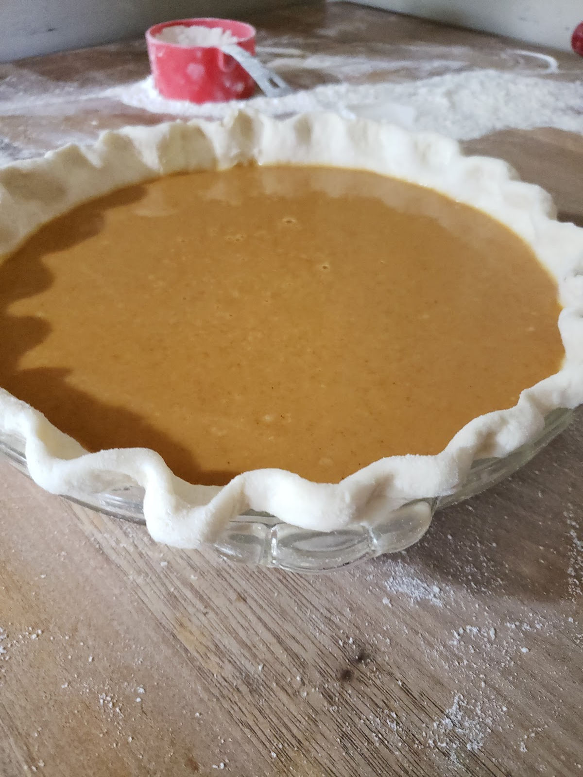 unbaked pumpkin pie on butcher block, red measuring cup with flour in background.