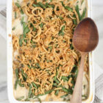 rectangle white baking dish with green bean casserole, wooden spoon on right edge of dish.