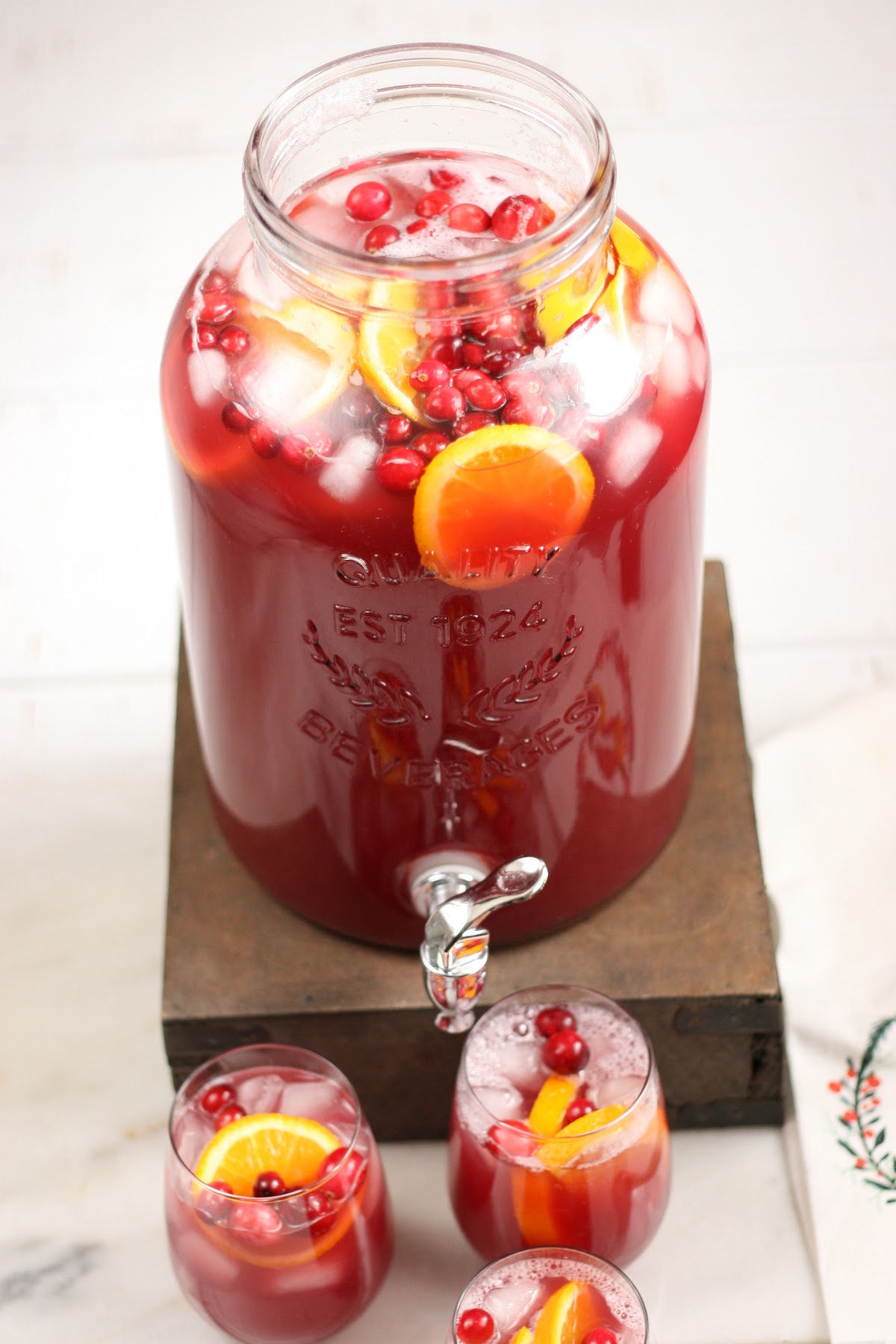 Clear drink dispenser of red punch, cranberries, orange slices, ice cubes.