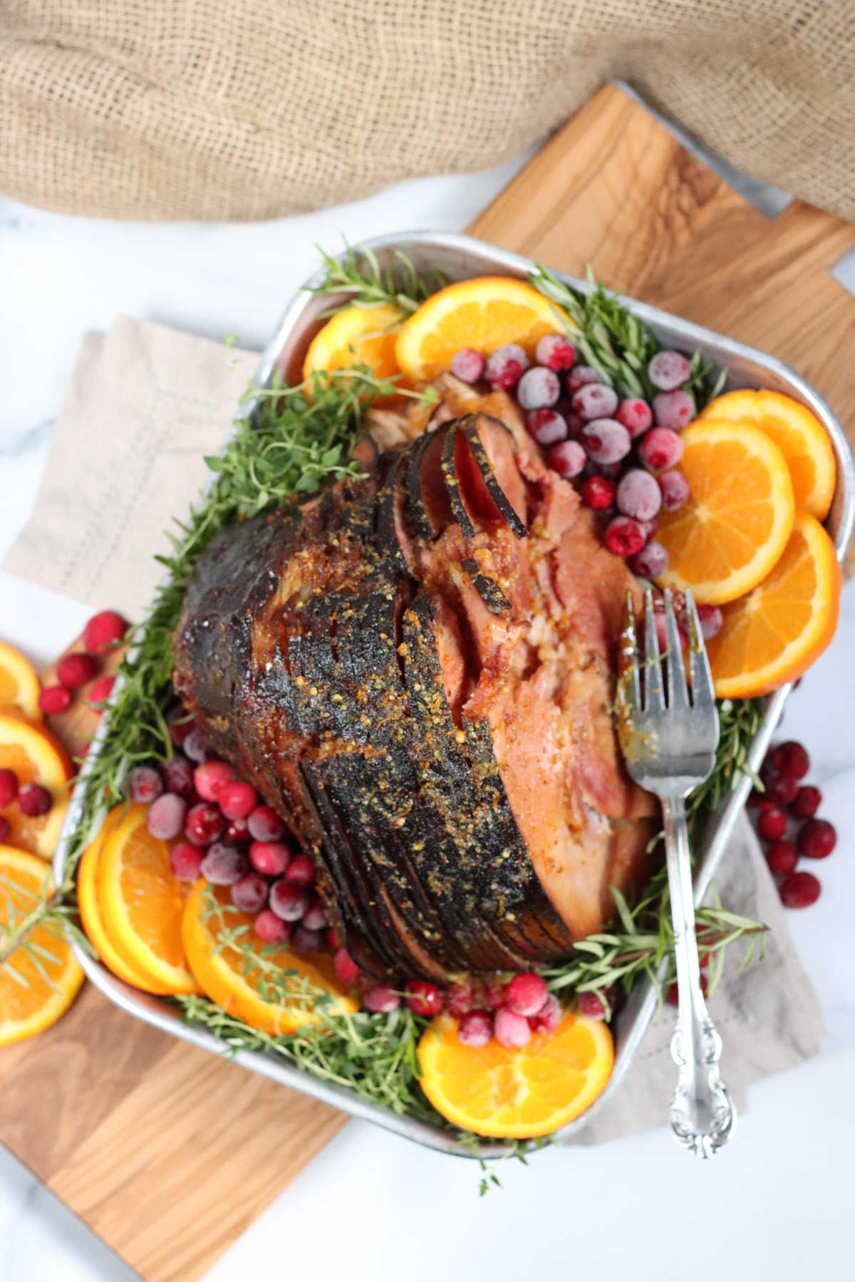 sliced ham in metal baking pan with orange slices, cranberries and herbs.