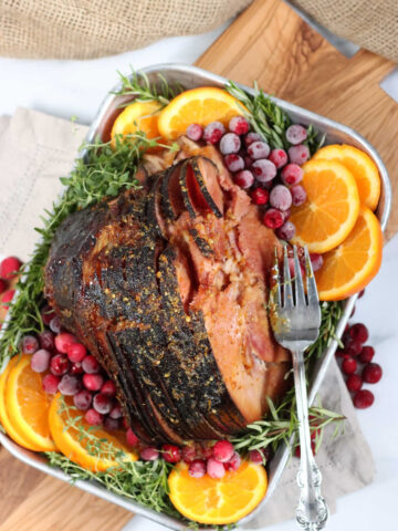 spiral cut ham in metal baking pan, orange slices, cranberries, fresh herbs, on cutting board.
