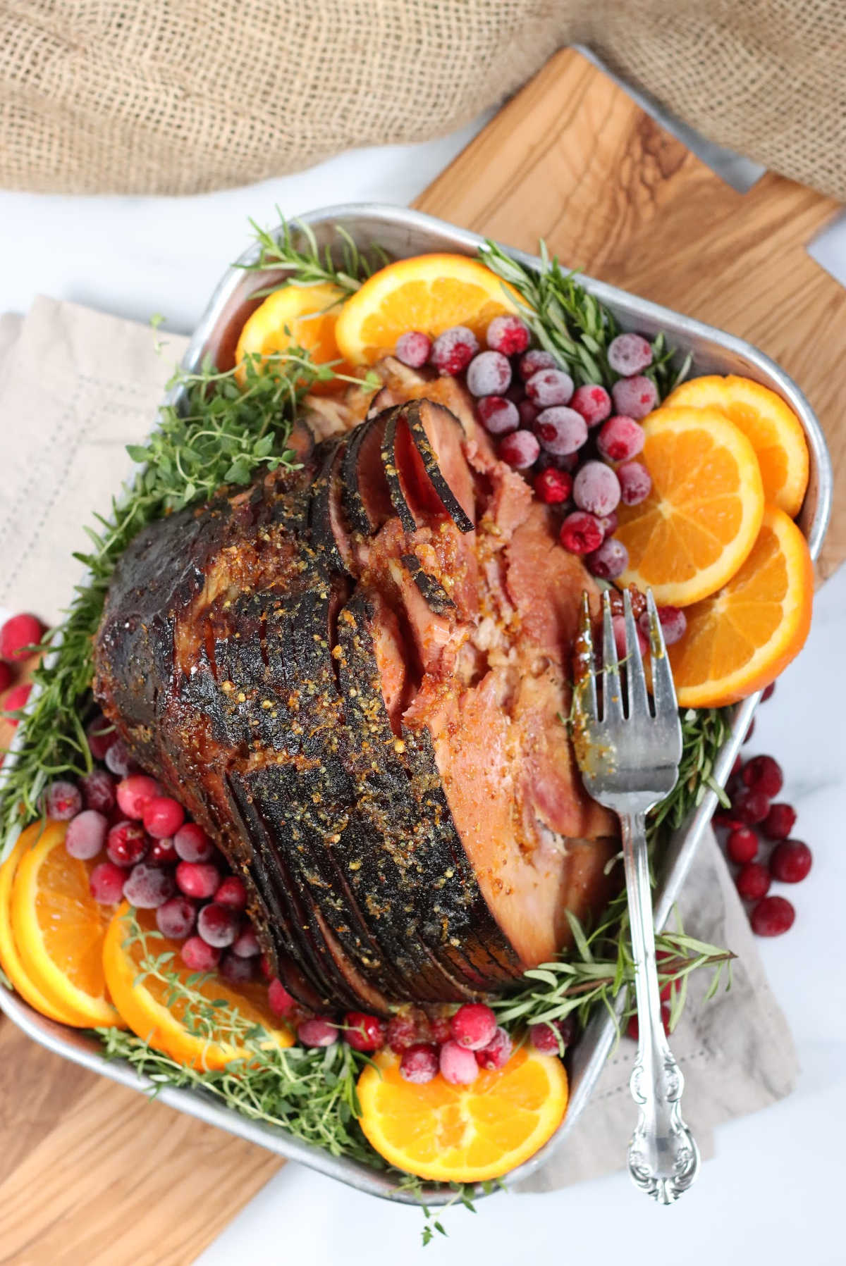 spiral sliced ham in a metal baking pan, surrounded by orange slices, cranberries and herbs.