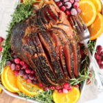 spiral cut ham in metal baking pan surrounded by orange slices, fresh cranberries, and herbs.