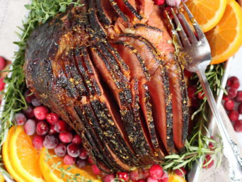 spiral cut ham in metal baking pan surrounded by orange slices, fresh cranberries, and herbs.