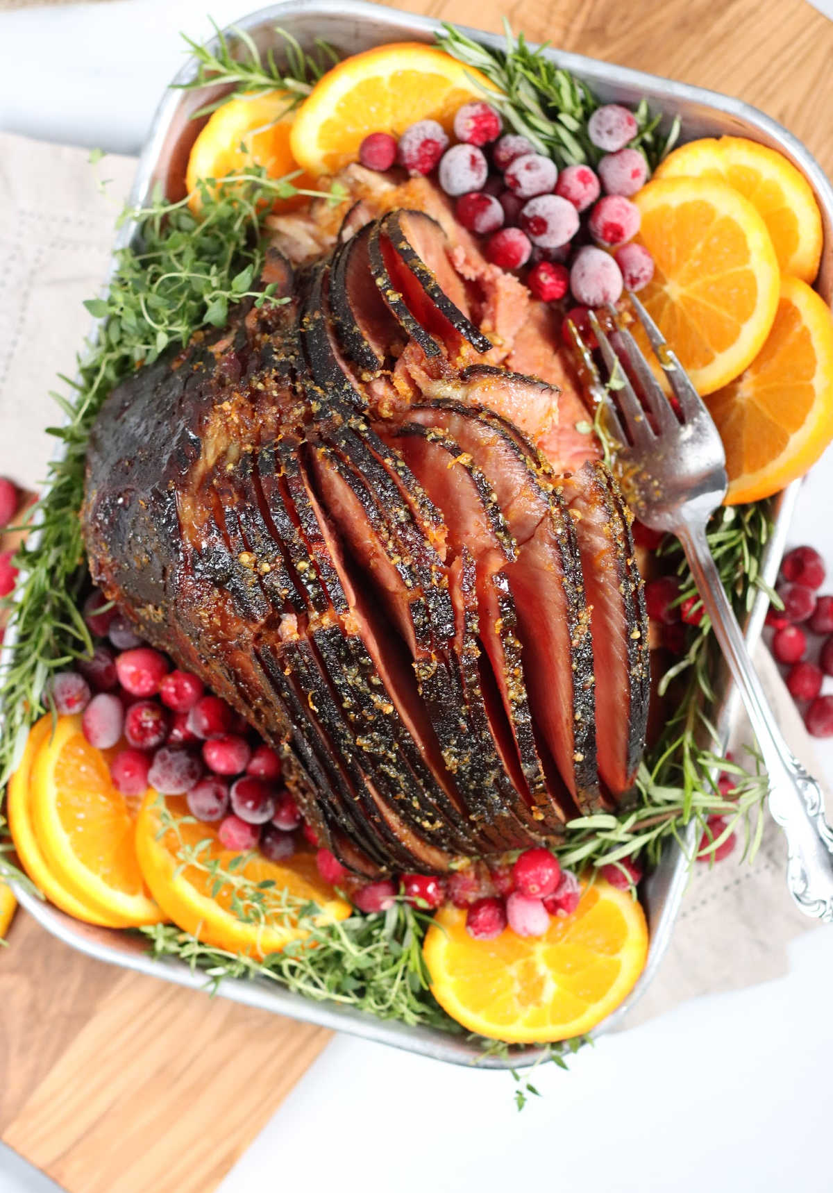 spiral cut ham in metal baking pan with orange slices, fresh cranberries, fresh herbs.