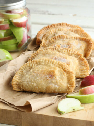 Four apple hand pies on wooden cutting board, slices of apples around them.