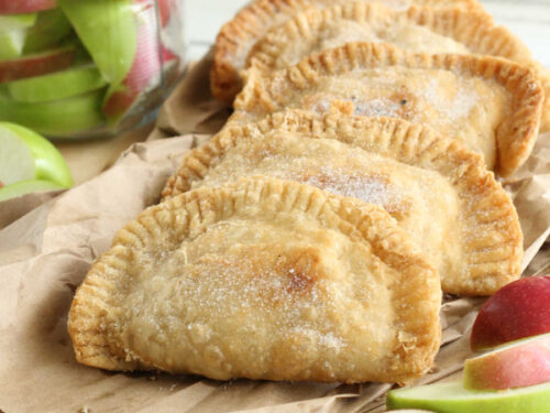 Four apple hand pies on wooden cutting board, slices of apples around them.
