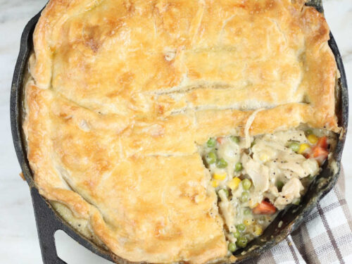 chicken pot pie with slice missing in dual handle cast iron skillet on taupe kitchen towel.