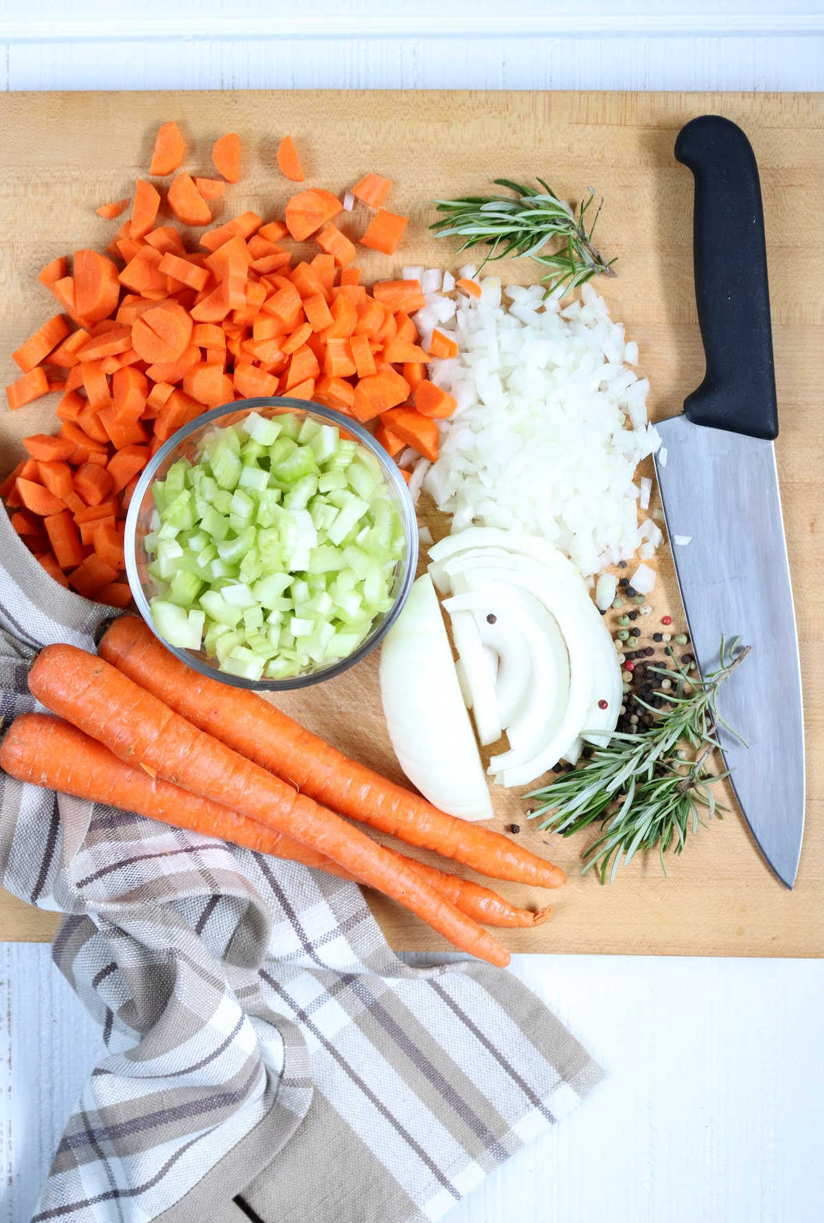 Chopped carrots, celery, onions and fresh rosemary for chicken pot pie on wooden cutting board with chef's knife.
