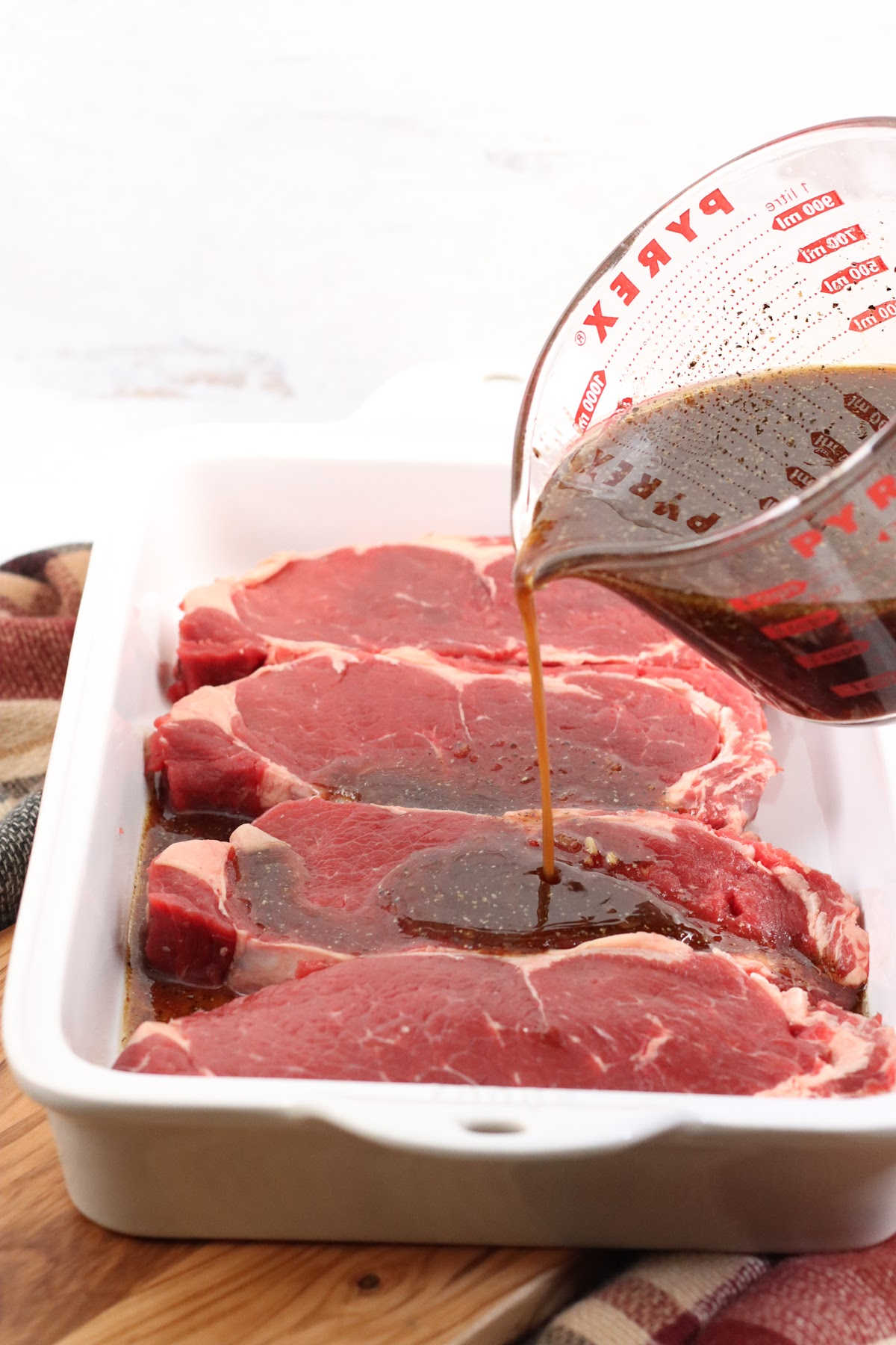Ribeye steaks in white ceramic baking dish, marinade being poured over top.