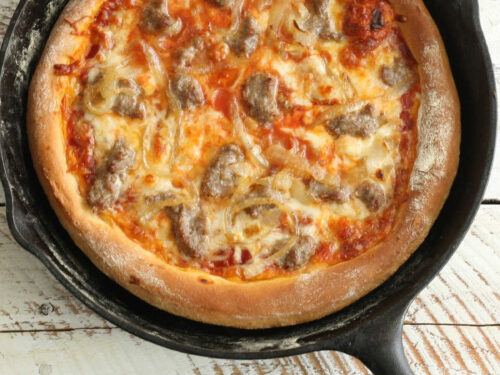 Homemade pizza in a cast iron skillet with golden cheese and sausage pieces.