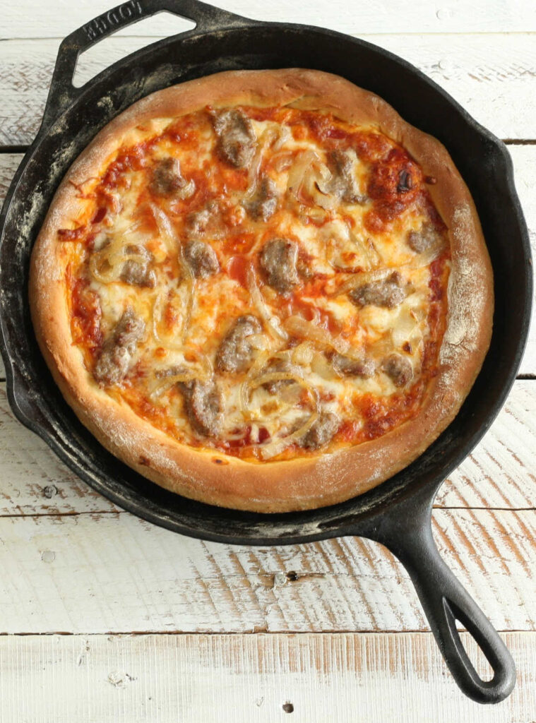 Homemade pizza in a cast iron skillet with golden cheese and sausage pieces.