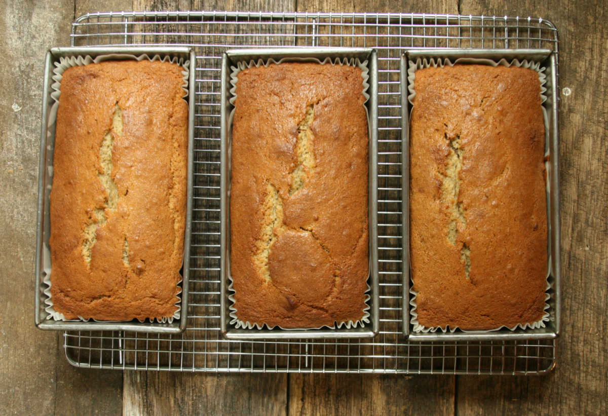 Three loaves of banana bread in metal loaf pans cooling on metal baking rack on reclaimed boards.
