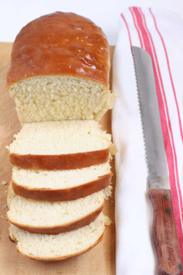 loaf of white bread partially sliced on wooden cutting board, wooden handle knife to right.
