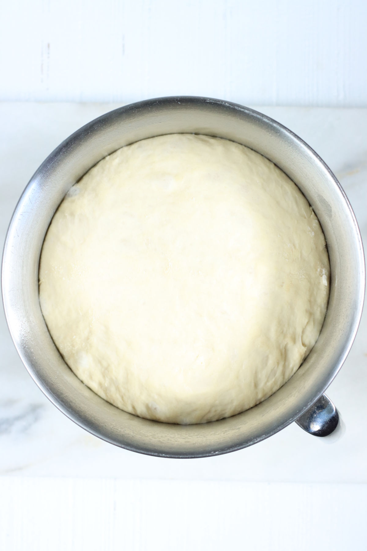 Metal mixing bowl with yeast bread dough rising on white marble.