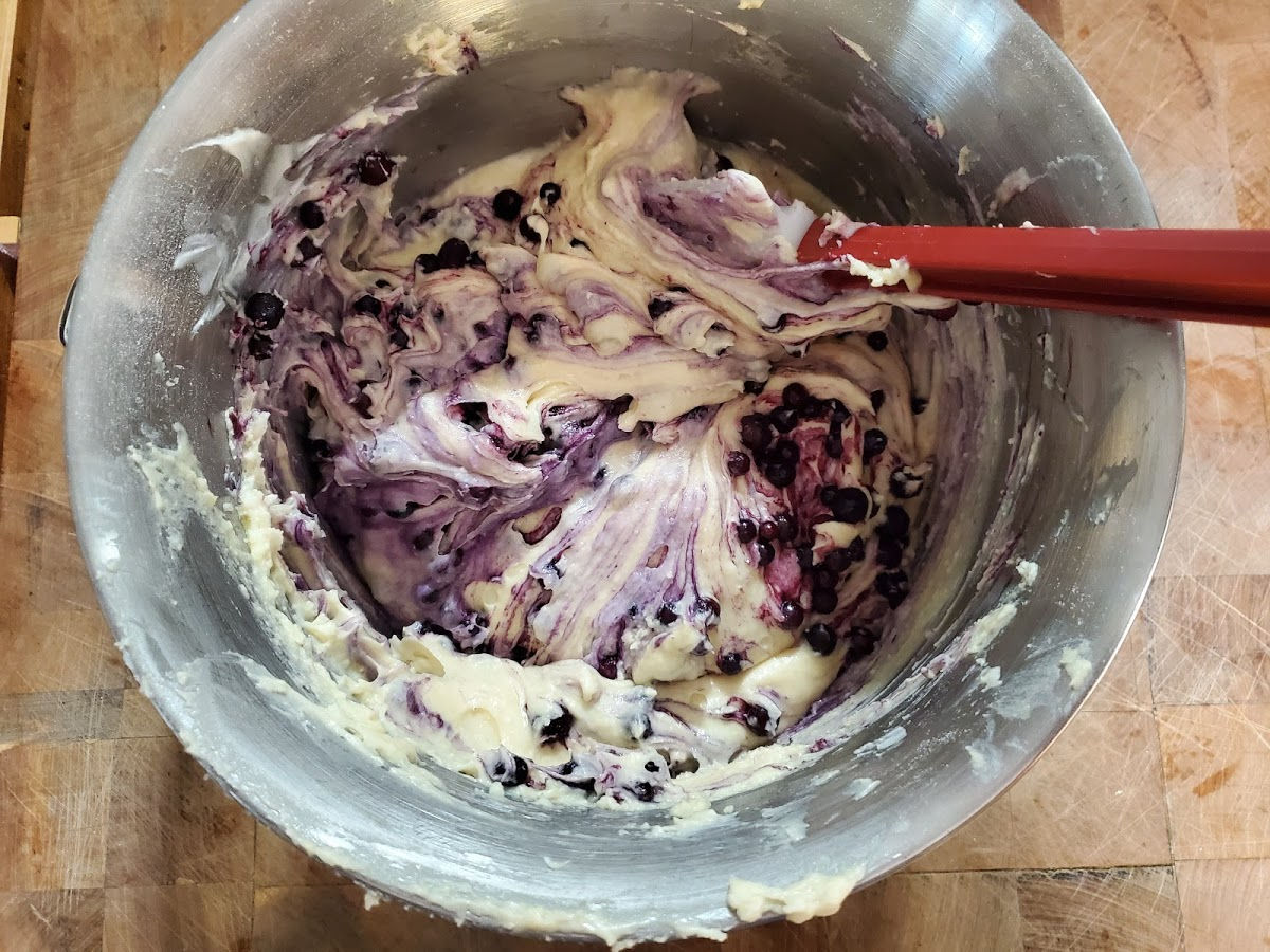Blueberry muffin batter in metal mixing bowl with deep red handle rubber spatula.