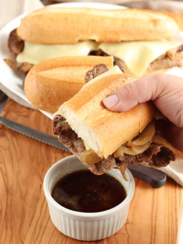 Half of French dip in a hand dipping into small white bowl of au jus.