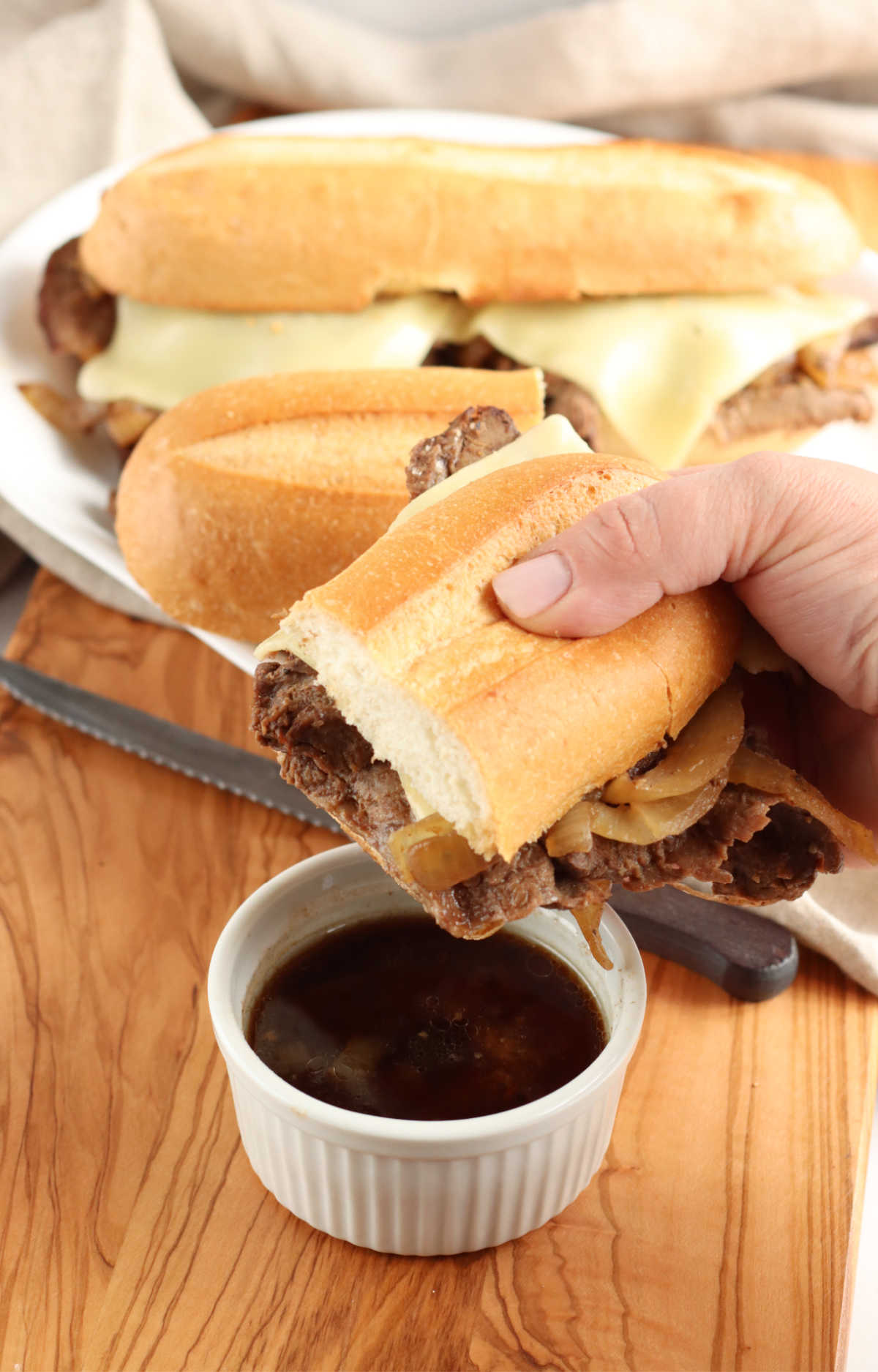 Hand holding French dip sandwich dipping into small white bowl of au jus.