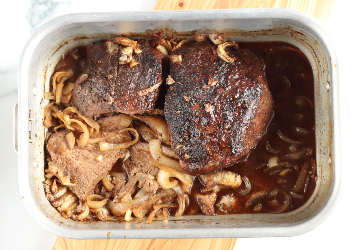 Two chuck beef roasts in metal roasting pan with caramelized onions and au jus.