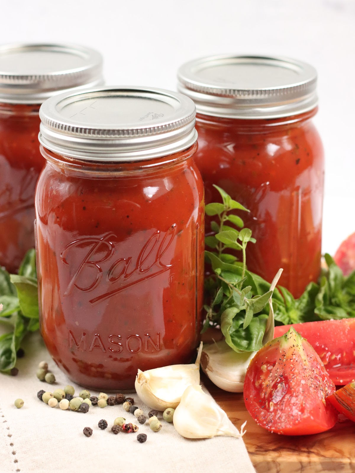 Marinara sauce in Mason jars on wooden cutting board, quarters of tomatoes, garlic cloves and fresh herbs.