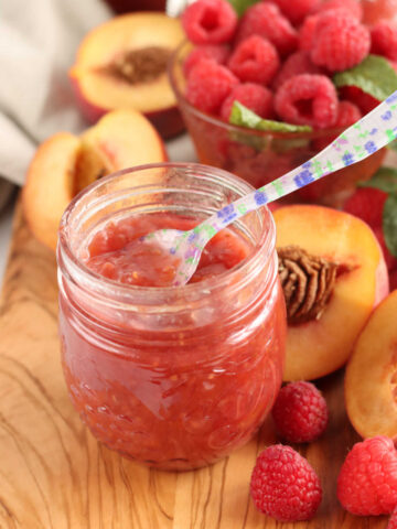 Jar of raspberry peach jam on wooden cutting board, floral spoon in jar.